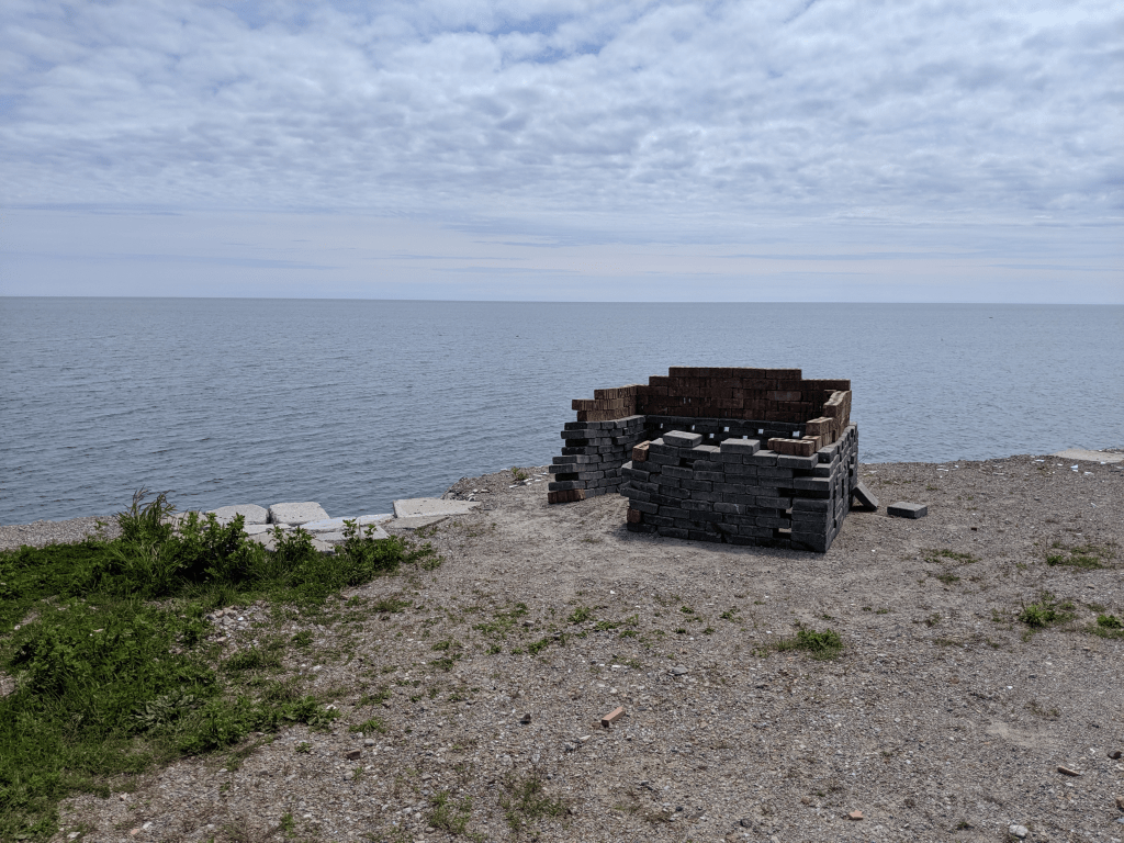A photo taken standing on the shoreline of Lake Ontario, at Tommy Thompson Park (Also known as the Leslie Spit). It is a cloudy summer day. In the foreground is a small, half built shelter made of grey bricks on the bottom, and red bricks for the top half. The left corner closest to the viewer has a gap big enough for a person to fit through. There is no roof.